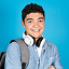 Smiling young man with short dark hair wearing a white sweatshirt and white headphones around his neck against a blue background.